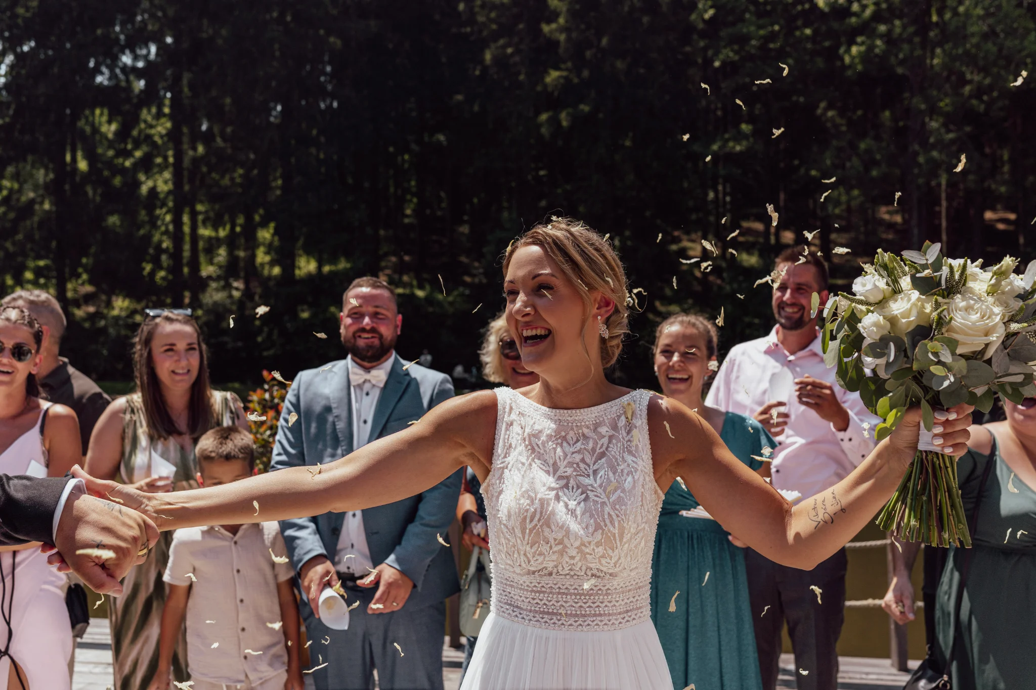 Smiling bride holding her husband's hand, carrying a bridal bouquet with flower petals in her hair.