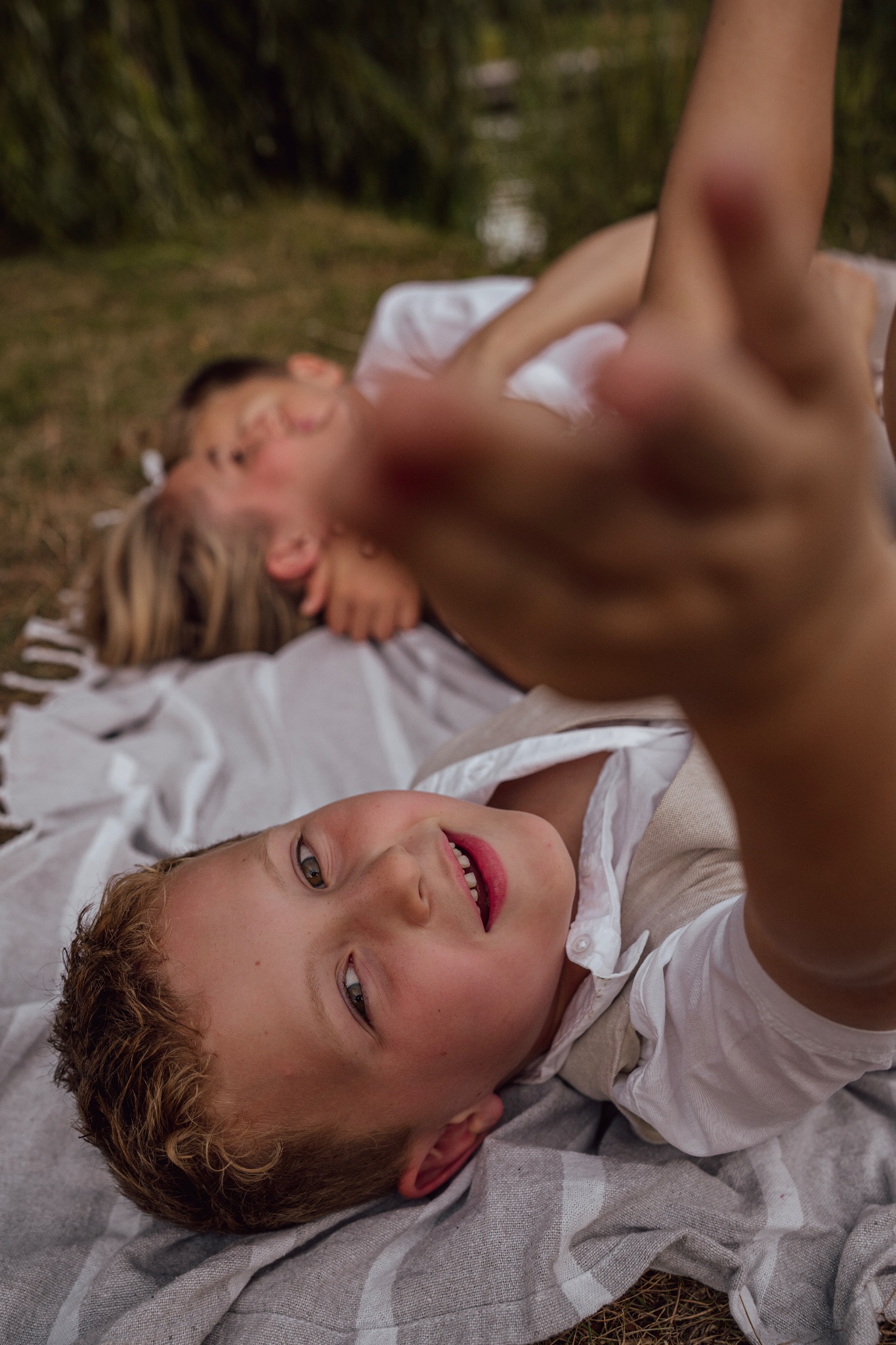 Little boy lying on a blanket and pointing at the camera, with his mother and brother behind him.