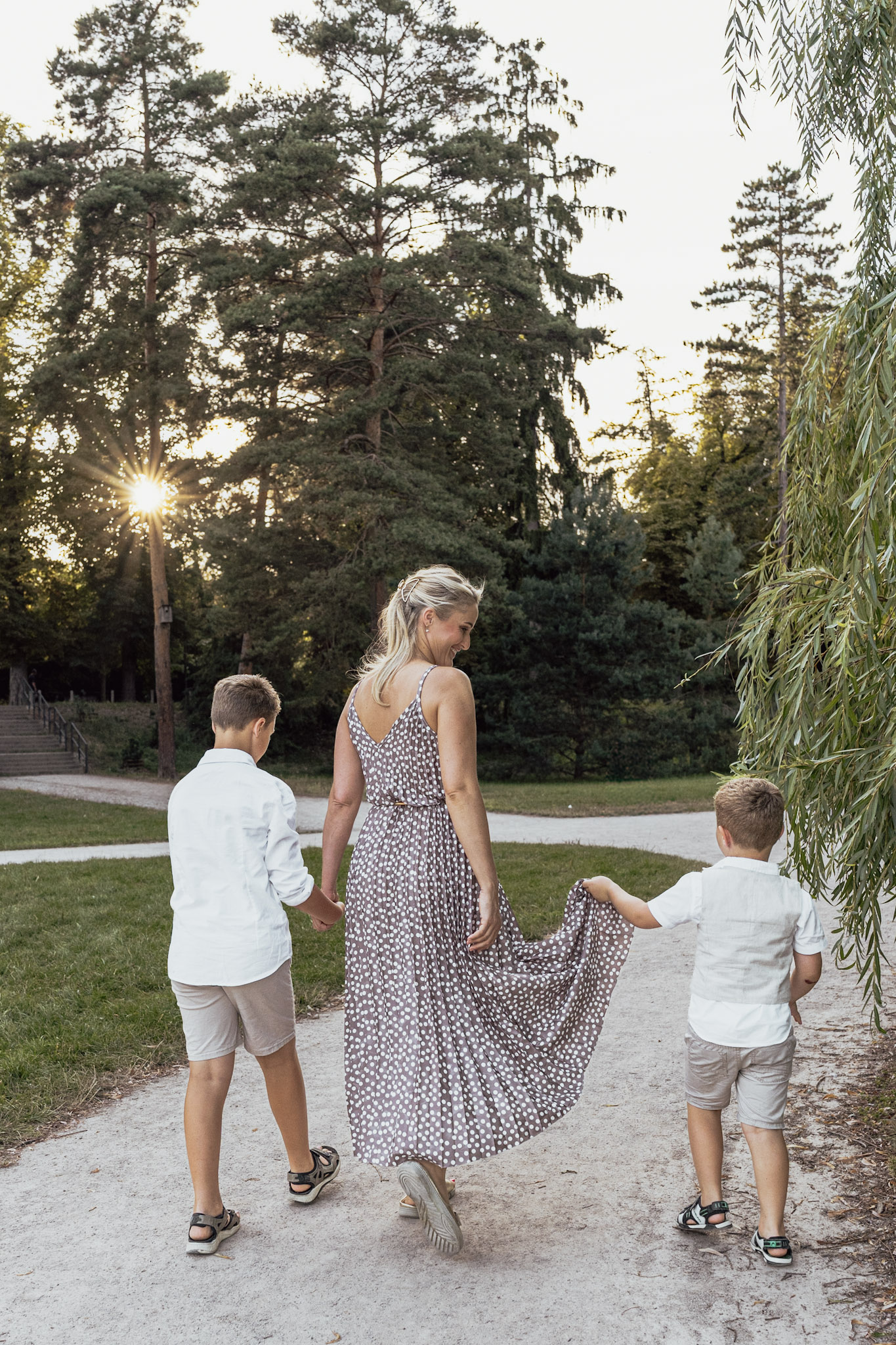 Mother walking through a park holding both of her sons’ hands, photographed from behind.